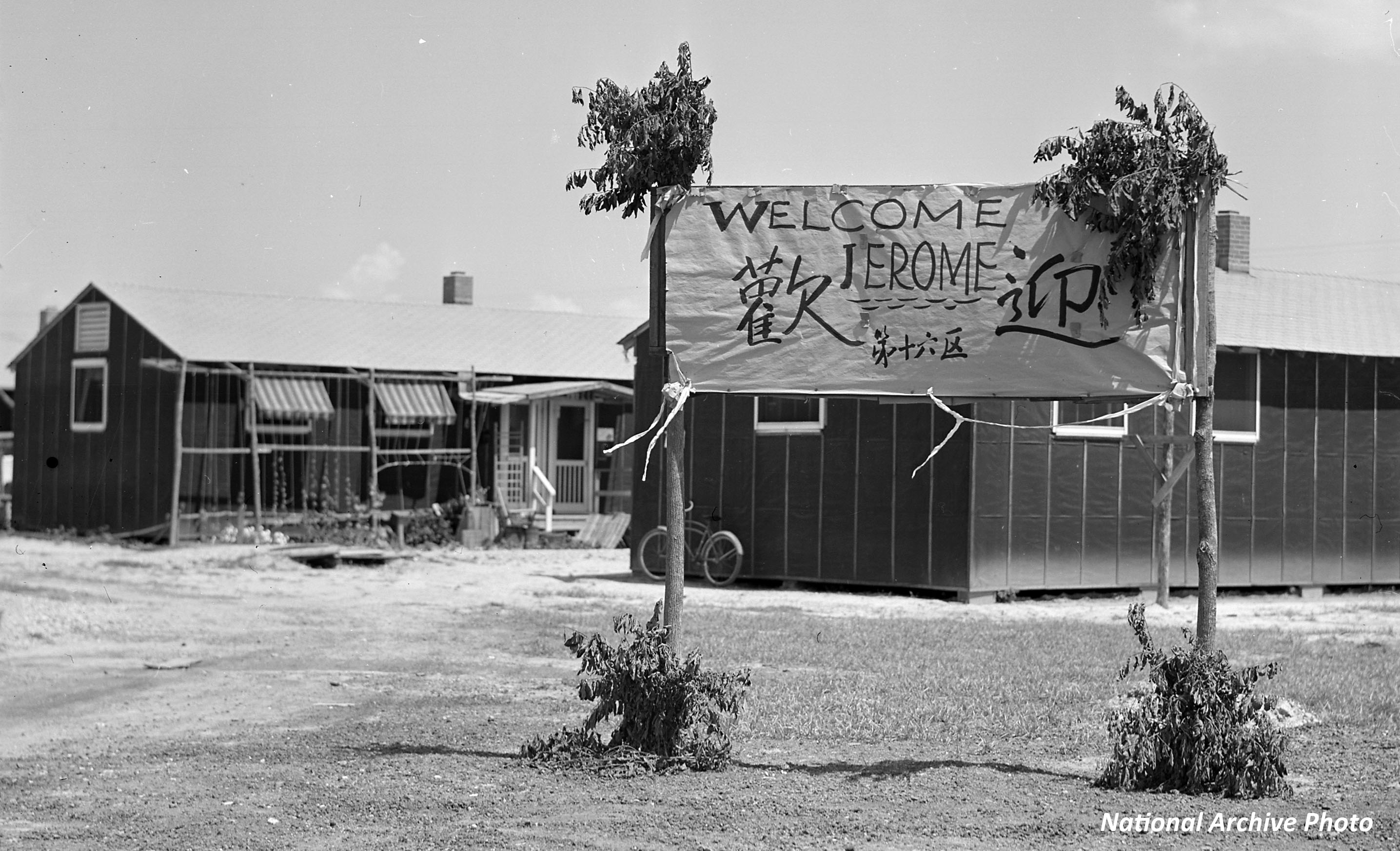 Two barracks at Rohwer Concentration Camp.  In front of the barracks are two poles decorated with leaves, so they look like trees.  Between the trees is a banner that states "Welcome Jerome"